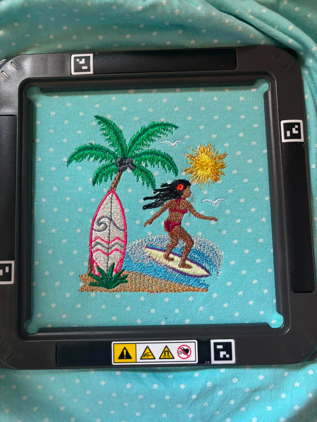 Motif de broderie Surfeuse disponible au format 10x10. Décor tropical avec palmier, planche de surf, océan et soleil, idéal pour une ambiance estivale.