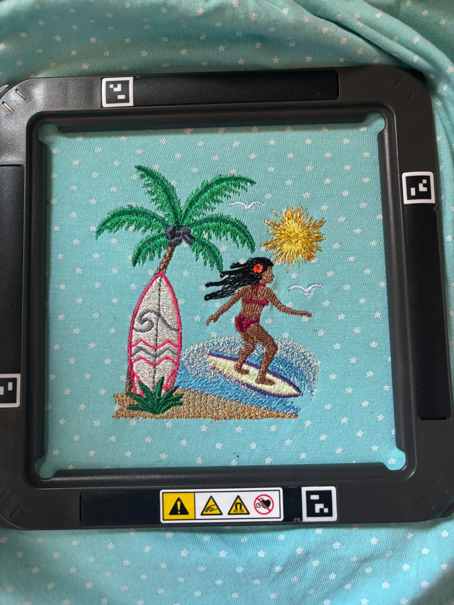 Motif de broderie Surfeuse disponible au format 10x10. Décor tropical avec palmier, planche de surf, océan et soleil, idéal pour une ambiance estivale.