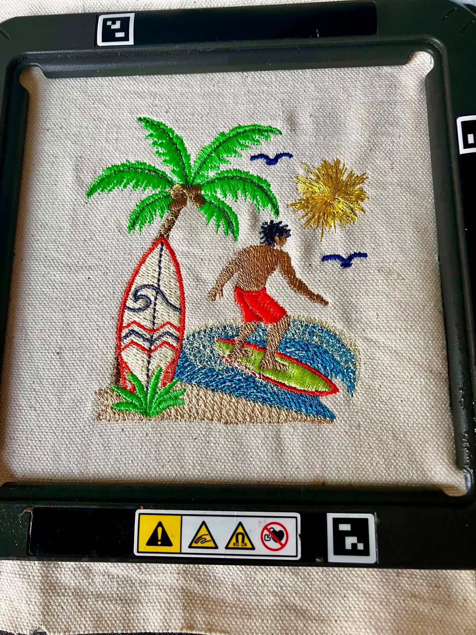 Motif de broderie Surfeur disponible au format 10x10. Décor tropical avec palmier, planche de surf, océan et soleil, idéal pour une ambiance estivale.
