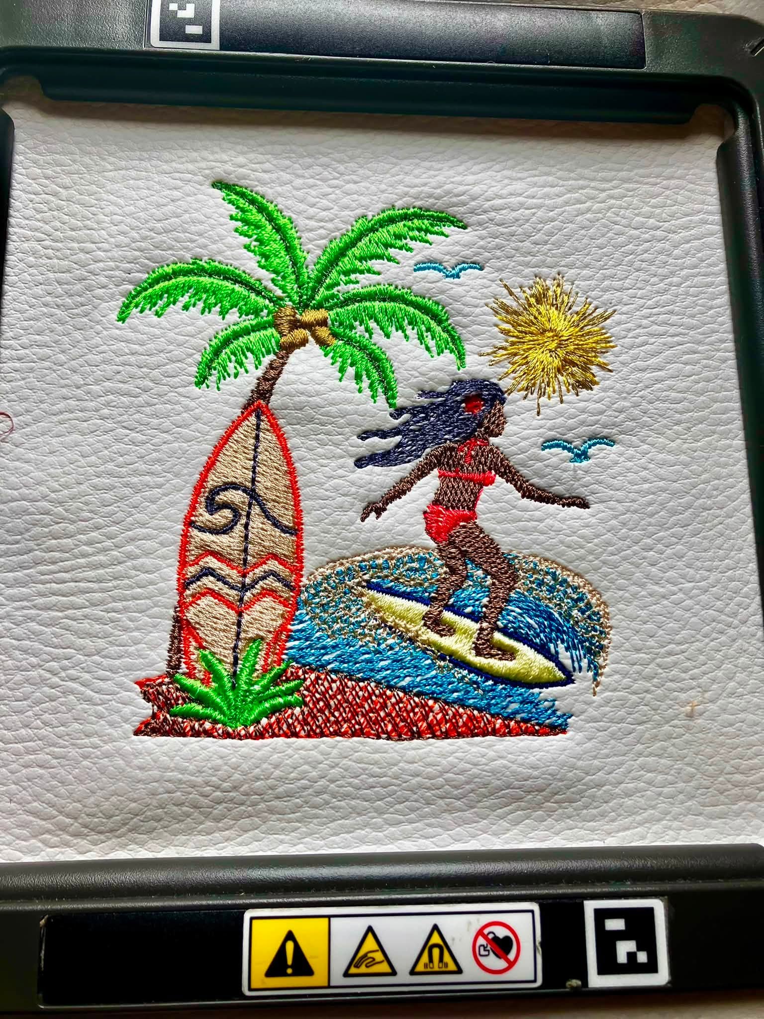 Motif de broderie Surfeuse disponible au format 10x10. Décor tropical avec palmier, planche de surf, océan et soleil, idéal pour une ambiance estivale.
