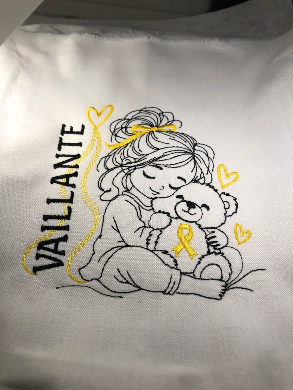 La Petite Vaillante, petite fille serrant un ourson avec ruban doré, cœurs et message VAILLANTE – motif broderie Septembre en Or